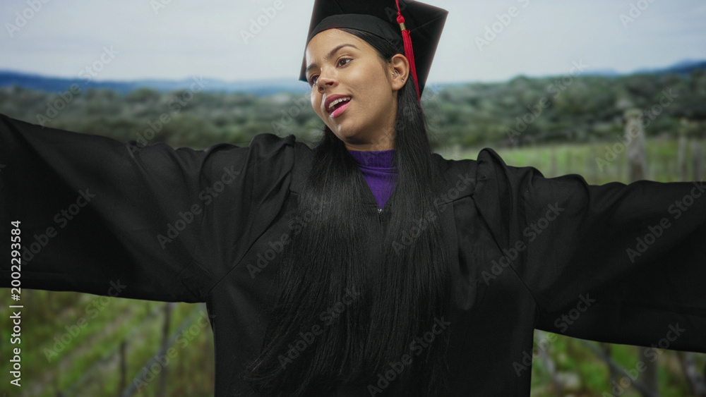 Obraz premium Young latina woman in black graduation gown and mortarboard with red tassel spreads arms in green countryside field; academic achievement pride.