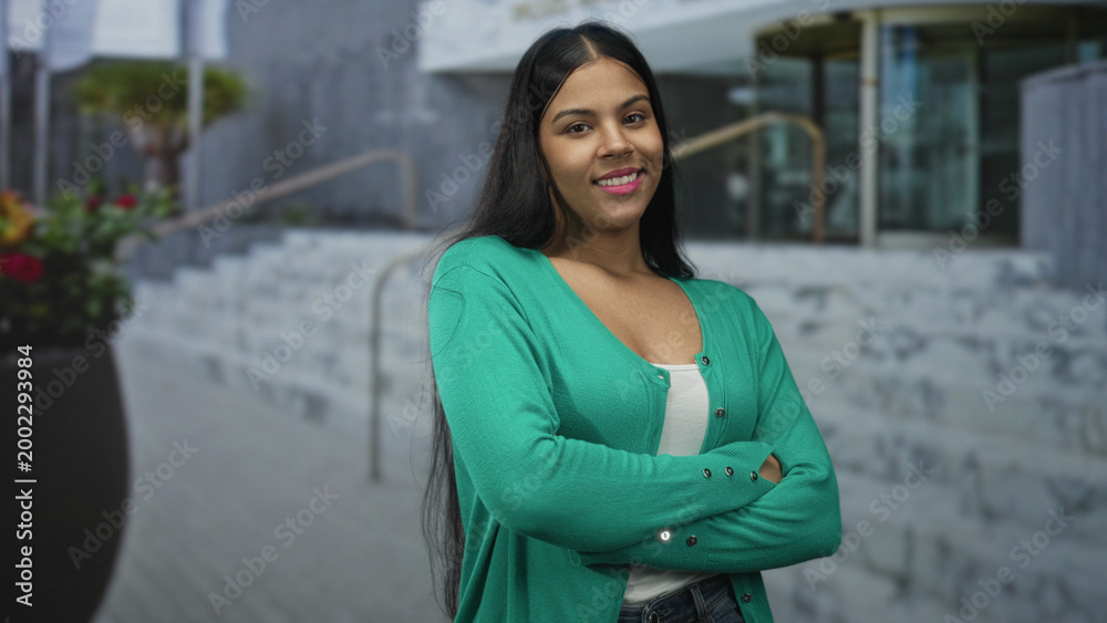 Fototapeta premium Woman stands with arms crossed in green cardigan at modern building entrance; confidence empowerment.