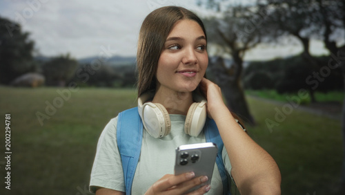 Quadro su tela Teenage girl holds smartphone in sunlit forest clearing while touching hair and looking aside; curiosity exploration discovery wonder