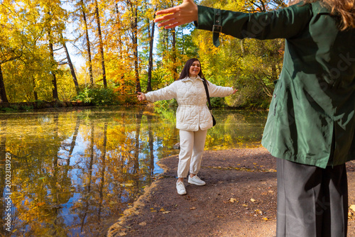 Woman in white jacket standing with open arms near pond in autumn park. Happy girl greeting friend in forest. Seasonal weekend activity, recreation in nature and friendship concept.