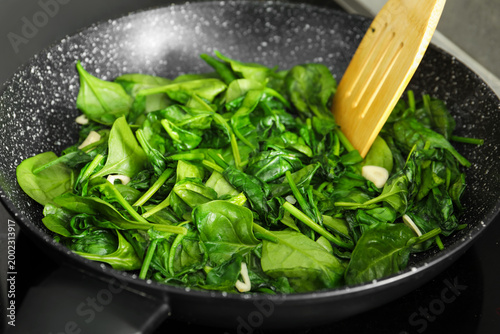 Photos Stirring spinach and garlic with wooden spatula in frying pan, closeup