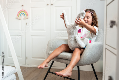Happy little girl hugging stuffed bunny on chair at home