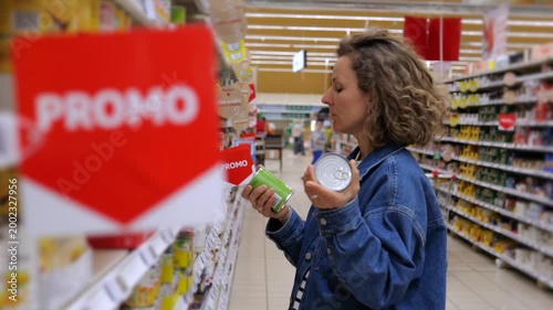 Frugal female shopper choosing canned food from a shelf in a supermarket aisle during a promotion, carefully comparing prices and products to save money on her grocery bill