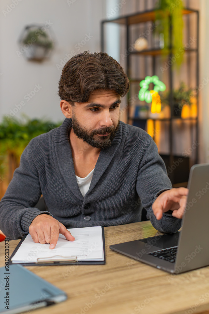 Obraz premium Indian man at home reviews paper reports compares numbers with laptop writes notes focused. Arabian guy at desk checks rows and figures on sheet reads data finds match does careful analysis calm mood