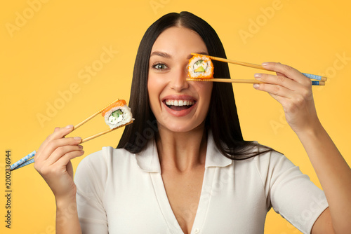 Fotografie Emotional young woman holding sushi rolls with chopsticks in each hand, covering
