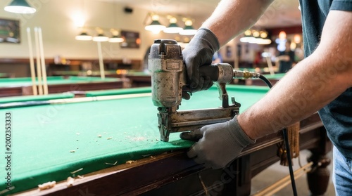 Worker using a pneumatic staple gun to attach green felt to the edge of a pool table