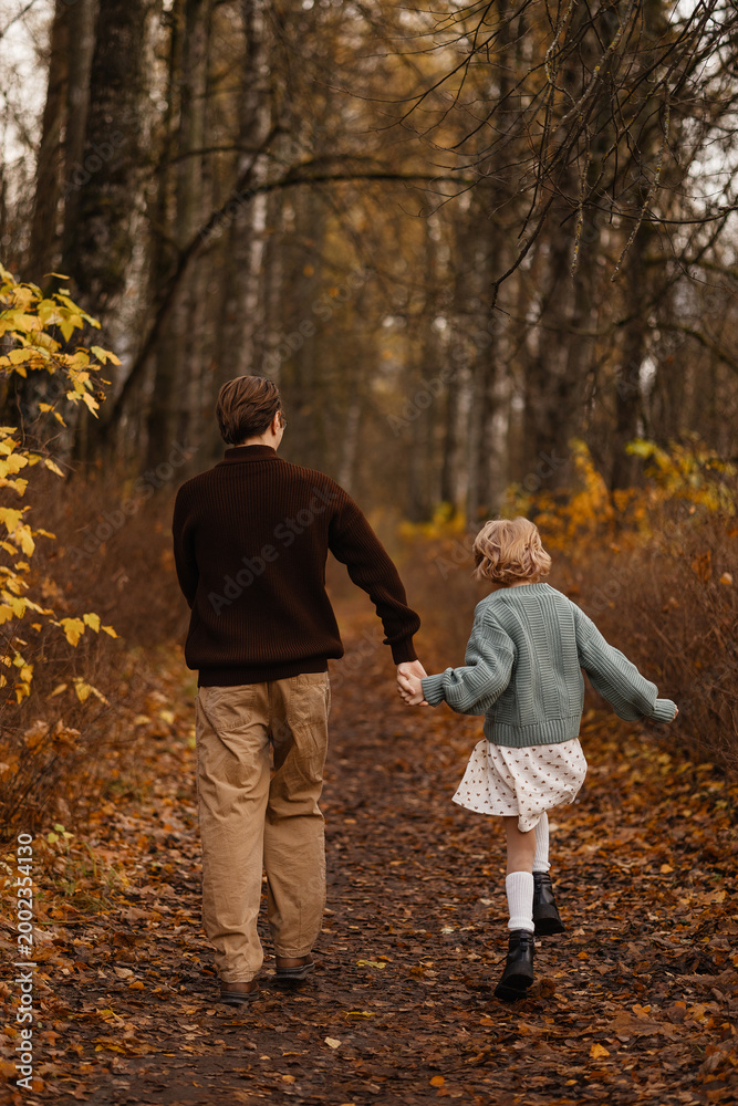 Fototapeta premium Back View Father And Daughter Walking Hand In Hand Down Leafy Trail Through Autumn Woods, Peaceful Reflective Scene With Golden Bushes, Slow Pace And Warm Seasonal Tones Evoking Journey