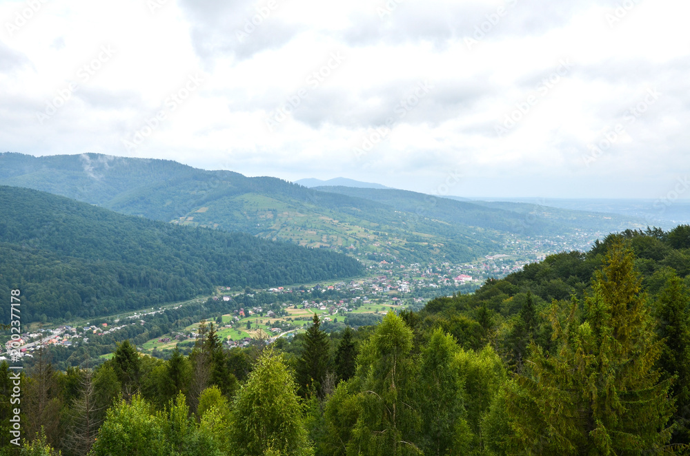Fototapeta premium Panoramic green mountain valley with Yaremche village nestled among dense forest and rolling hills, showing rural landscape, misty atmosphere and cloudy sky. Carpathian Mountains, Ukraine