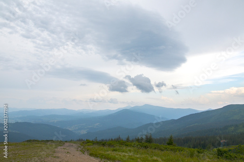 Wide panoramic view of layered blue mountains and a grassy foreground beneath dramatic cloud formations, conveying calm, solitude and expansive natural beauty. Carpathian Mountains, Ukraine