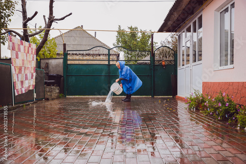 Portrait of an elderly woman in a blue raincoat pouring water from a bucket in the yard.