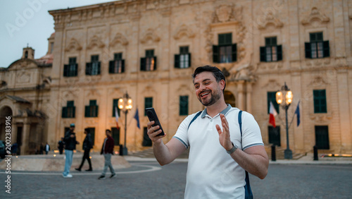 Fotografie Man using smartphone on a street in Malta during evening time with historic buil