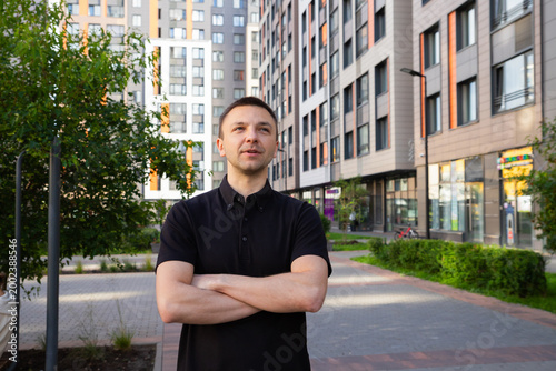 Young man entrepreneur standing with arms crossed in front of modern apartment building. Successful businessman looking away. City lifestyle, apartment investment and urban development.