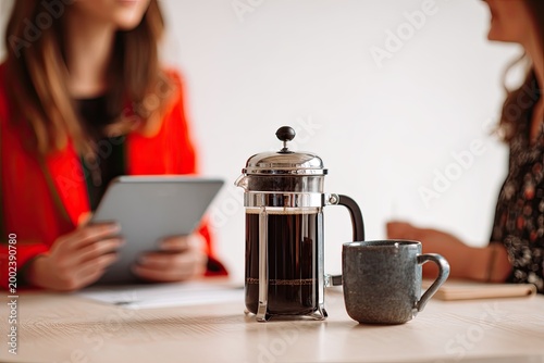 Two individuals engage in a meeting or discussion while enjoying coffee brewed in a French press, with a tablet resting on a wooden table.