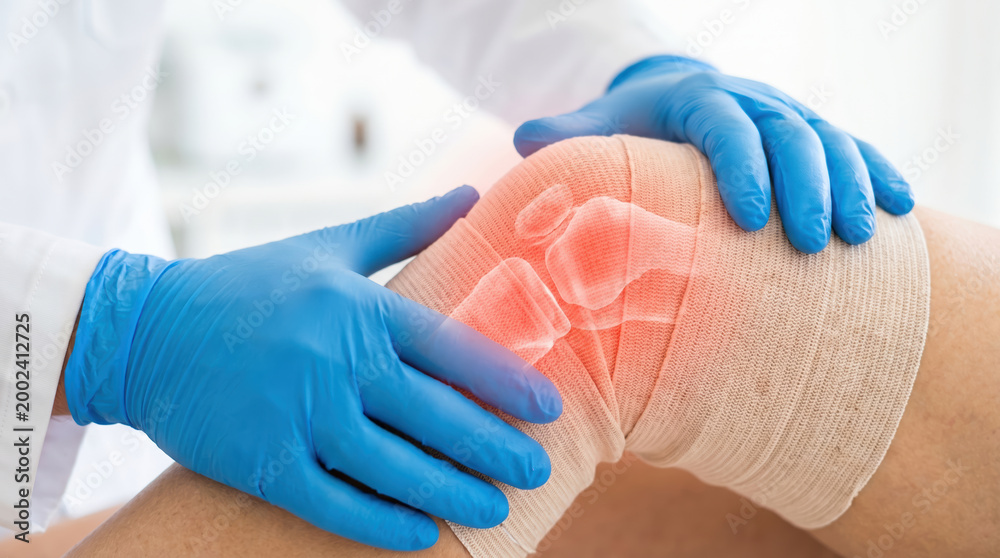 Fototapeta premium A medical professional provides care for a patient's injured knee. The doctor examines the knee while applying bandages. The setting shows a clean clinic with medical supplies nearby