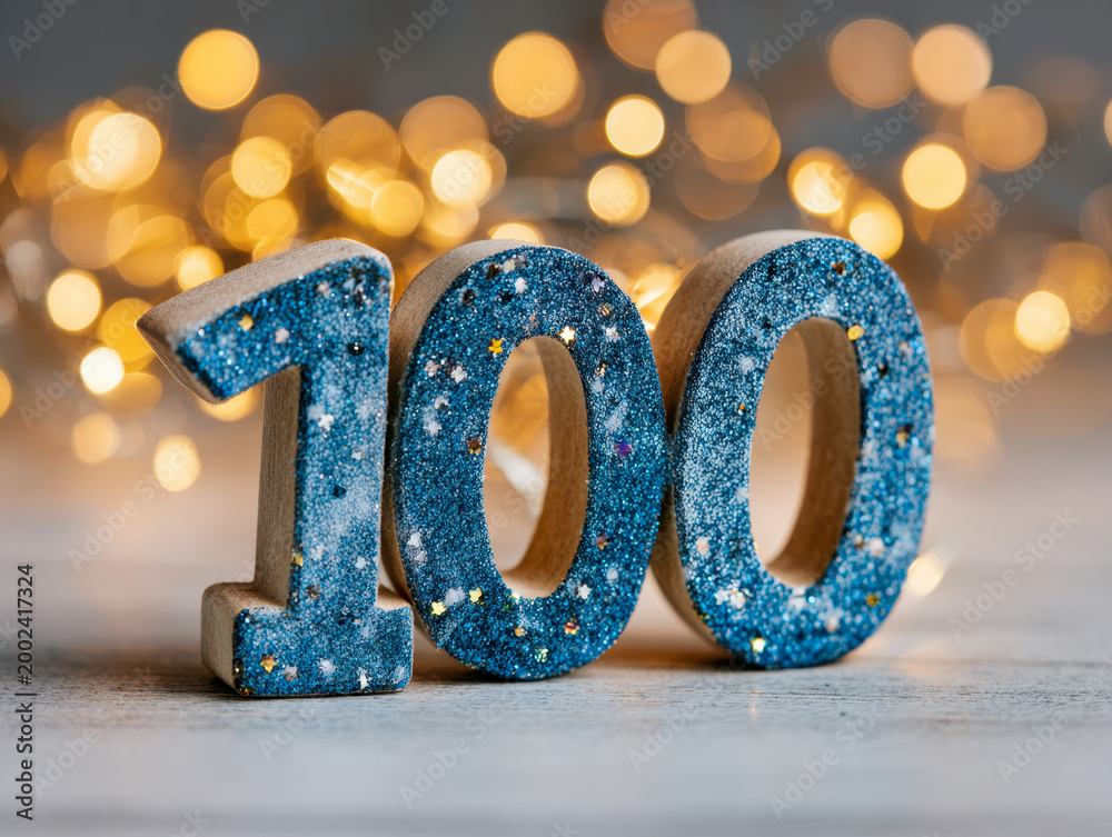 Fototapeta premium Sparkling blue glitter decorative numbers forming one hundred with warm glowing bokeh lights in the background for celebration or milestone concepts