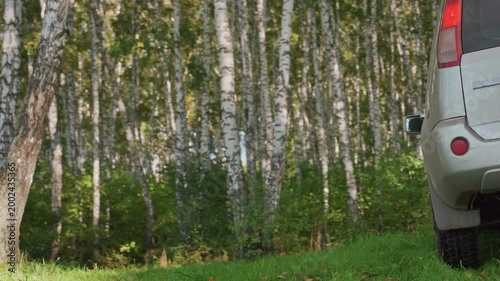 Woman camper exits muddy vehicle at birch forest edge, opens rear door and steps onto grassy clearing, sunlight through canopy, casual jacket, preparing gear for weekend camping escape