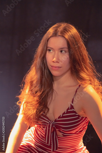 A confident young woman in a striped dress portrays a contemplative gaze, creating an emotive connection with the viewer against a soft dark backdrop that elevates her beauty.
