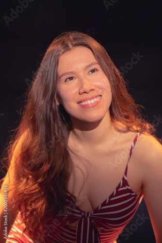 A charming young woman in a striped dress radiates pure joy with her smile, creating a connection with the viewer, captured in an artistic glow against a rich dark background.