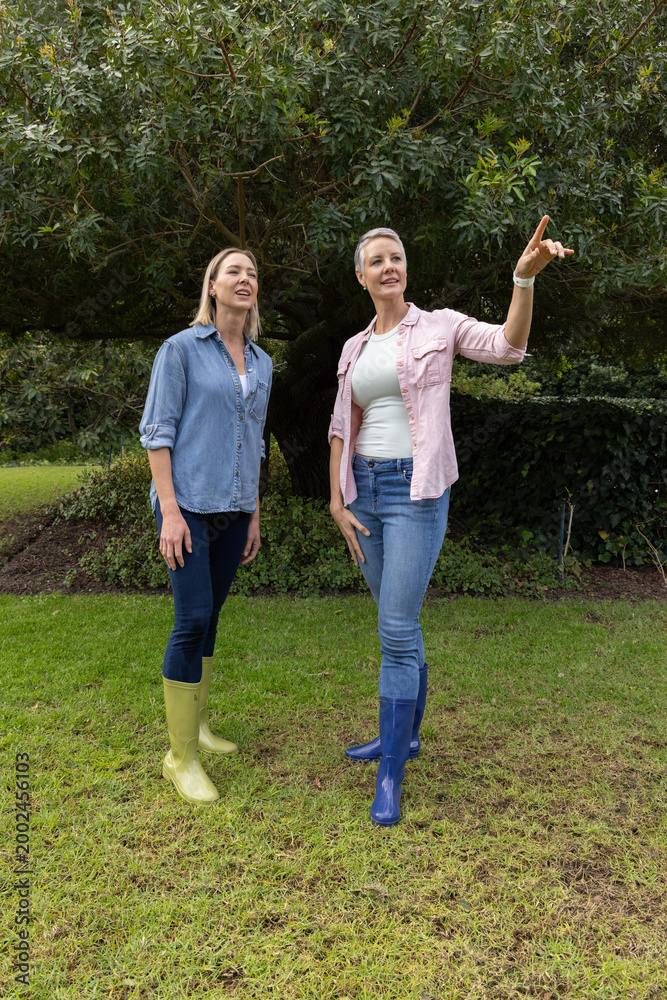 Obraz premium Two female friends standing on grass beneath tree pointing right, wearing Wellington boots, watch