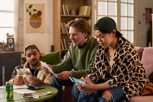 Фотография Multiethnic young adults reviewing bills, counting cash, using smartphone for budgeting at home