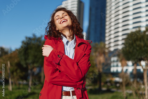 Fotografia Smiling young woman with curly brunette hair wearing a red trench coat stands in a sunlit urban park, enjoying a carefree moment outdoors