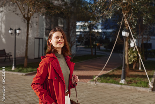 Foto Young woman in a red coat outdoors, smiling under afternoon light, walking in a