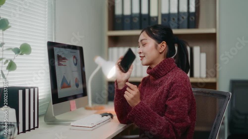 Young Asian woman working at her desk, having a business conversation on her smartphone while looking at charts on her computer screen