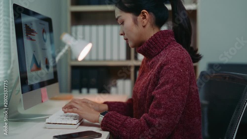 Professional asian woman sitting at her desk, typing on the keyboard and looking at financial charts and graphs on her computer monitor
