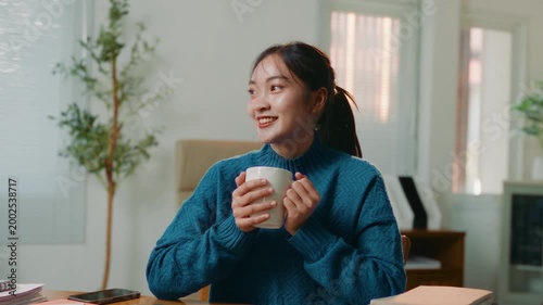 Young Asian woman in a cozy sweater smiling while enjoying a warm cup of coffee or tea during a break from work in her home office
