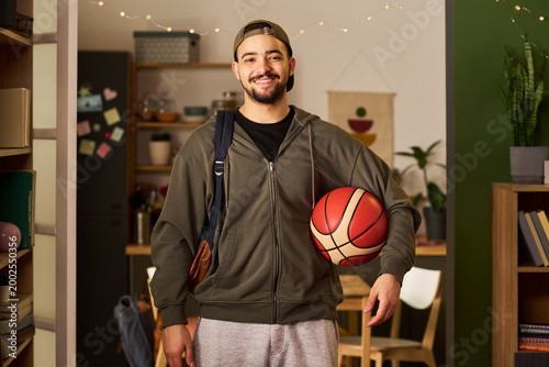 Fotografía Portrait of young adult biracial man carrying basketball and bag, smiling at camera in home interior, representing student lifestyle and active routine