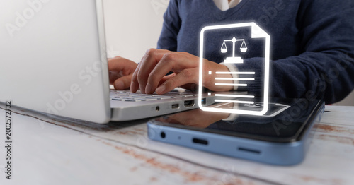 A person types on their laptop at a desk. Their fingers move with concentration across the keyboard. A smartphone rests next to the open laptop, displaying a legal document icon.
