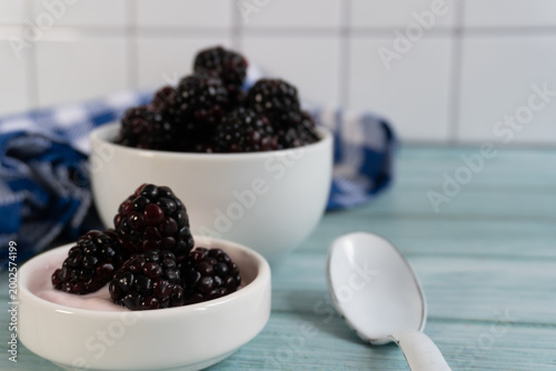 Fresh blackberries rest in white bowls on a wooden surface. Blue and white cloth adds soft contrast to the scene. Spoon lies beside the bowls, ready for use
