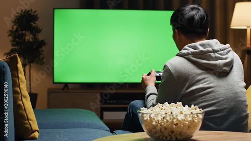A person is sitting on a sofa, playing a video game on a large television screen with a green background, with a bowl of popcorn nearby