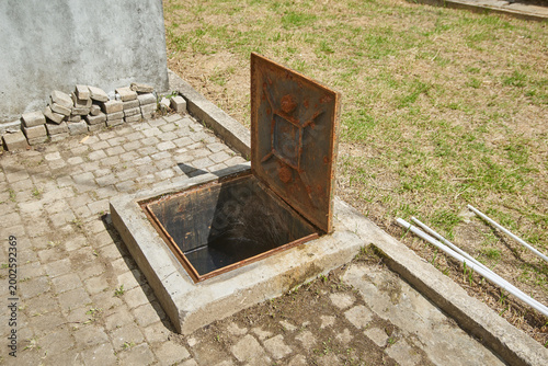 concrete underground tank with open rusty metal lid, dark water chamber surrounded by cobblestones, soft focus with copy space