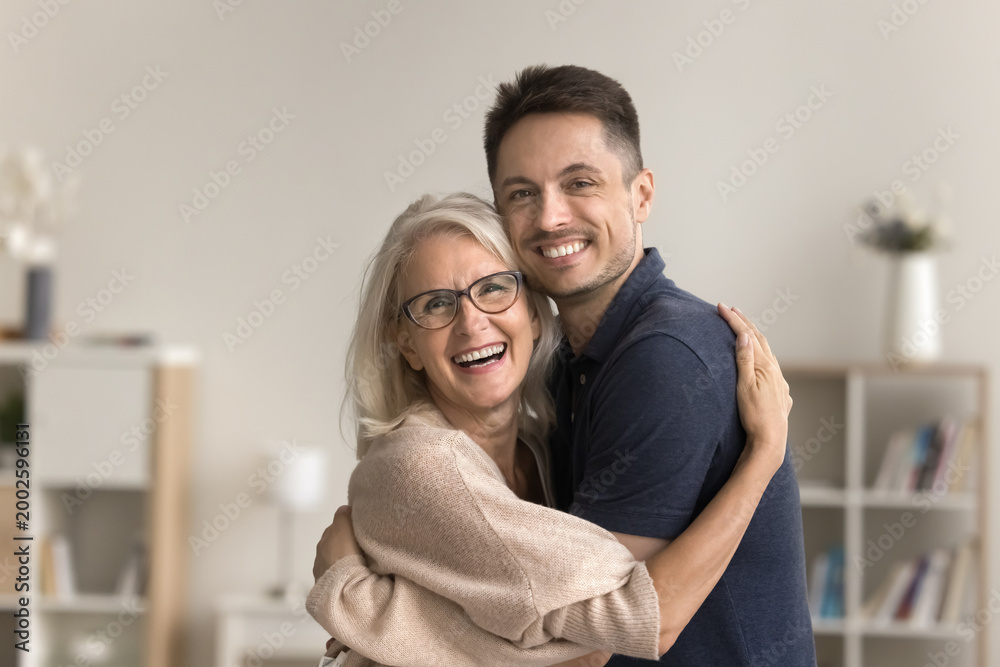Fototapeta premium Beautiful mature woman cuddling her young adult son, laughing, pose in domestic room, look for camera with positive, joyful expression. Family attachment, love, support. Happy Mothers Day celebration