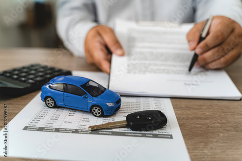 Hands reviewing car loan finance document contract with payment schedule table, blue toy car model and key on paperwork, calculator on desk, representing auto finance agreement analysis, vehicle lease