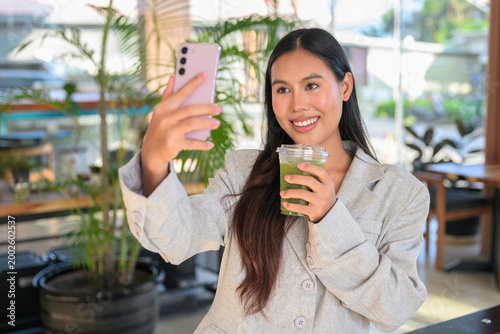 Smiling Thai woman taking a selfie and holding a matcha tea in a cafe, capturing a natural, professional lifestyle moment of a student.