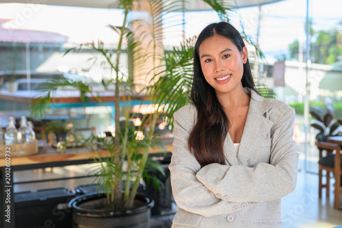 Smiling Thai woman wearing a blazer with arms crossed in a cafe, capturing a natural, professional lifestyle moment of a student.