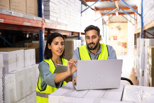 Photography Workers working in storage aisle, using laptop and digital tablet to review inventory data, check stock levels, coordinate logistics operations