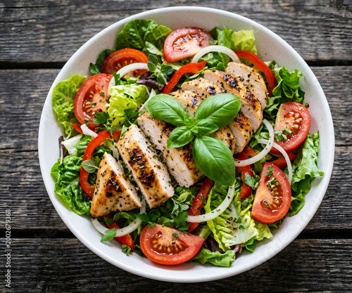 Grilled chicken salad with tomatoes lettuce onions and bell peppers on wooden table leafy greens white bowl light meal