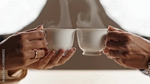 Two Pairs Of Hands Holding Steaming White Tea Cups With Gold Trim In A Warm Light Indoors