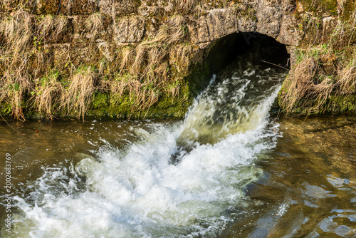 Water flows from a stone drain into a river in a rural area during daylight hours near a natural landscape with grass and rocks