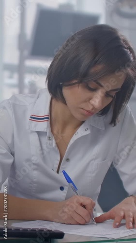 Focused female professional in white uniform writing notes at modern desk in bright office environment, concentrating on paperwork and administrative documentation tasks