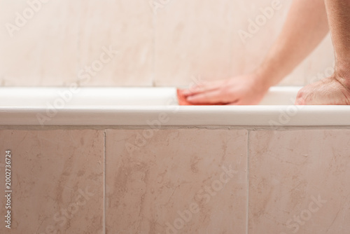 Closeup photo of man using polishing sandpaper sponge for bath cleaning. Sanding bathtub edge to prepare surface for new sealant