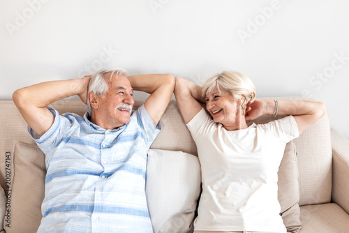 Fotografie Relaxed senior couple sitting on couch with hands behind heads, smiling elderly husband and wife enjoying retirement, leisure and comfort concept
