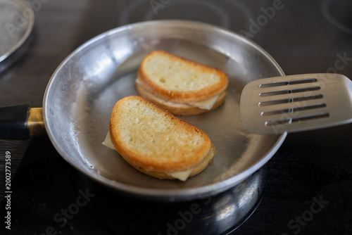 Grilled sandwiches sizzle in a frying pan. Golden-brown bread hints at perfect toasting. Metal spatula rests beside the pan. Close-up view highlights kitchen cooking scene