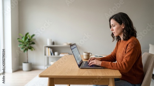 A woman wearing an orange sweater is working on her laptop at a wooden desk in a well-lit room, with a plant and bookshelf in the background