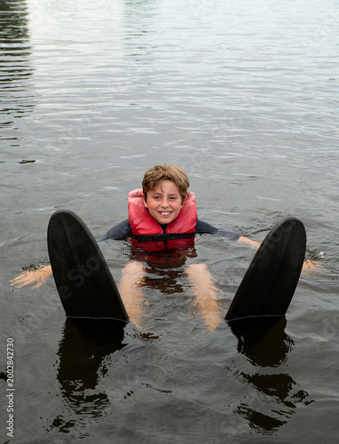 Obraz na płótnie Smiling Teenage Boy in Red Life Jacket Sitting on Water with Water Skis
