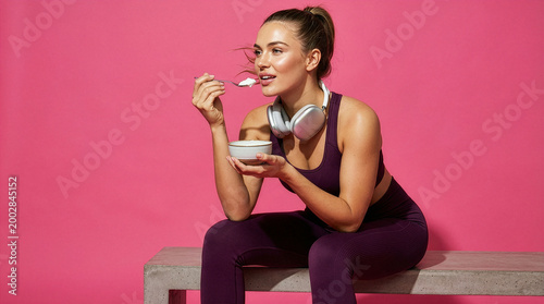 Fit young woman with headphones holding spoon and bowl on pink backdrop