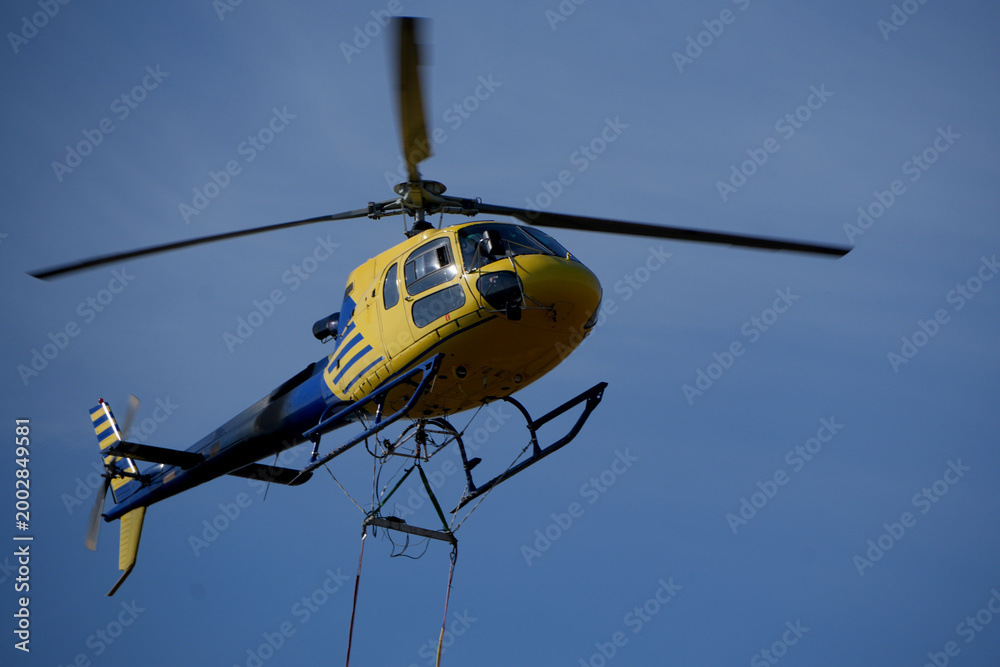 Fototapeta premium blue and yellow helicopter flying in a clear blue sky carrying equipment